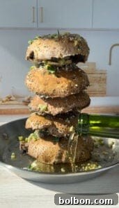 A stack of large, seasoned portobello mushrooms in a skillet, with olive oil being poured from a green bottle. The mushrooms are garnished with herbs and garlic, and the pan is sitting on a wooden surface. A minimal kitchen backdrop is visible.
