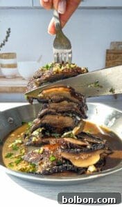 A fork and knife are slicing through a stack of cooked Portobello mushrooms, topped with chopped parsley and garlic, in a pan filled with sauce. The kitchen background is softly blurred.