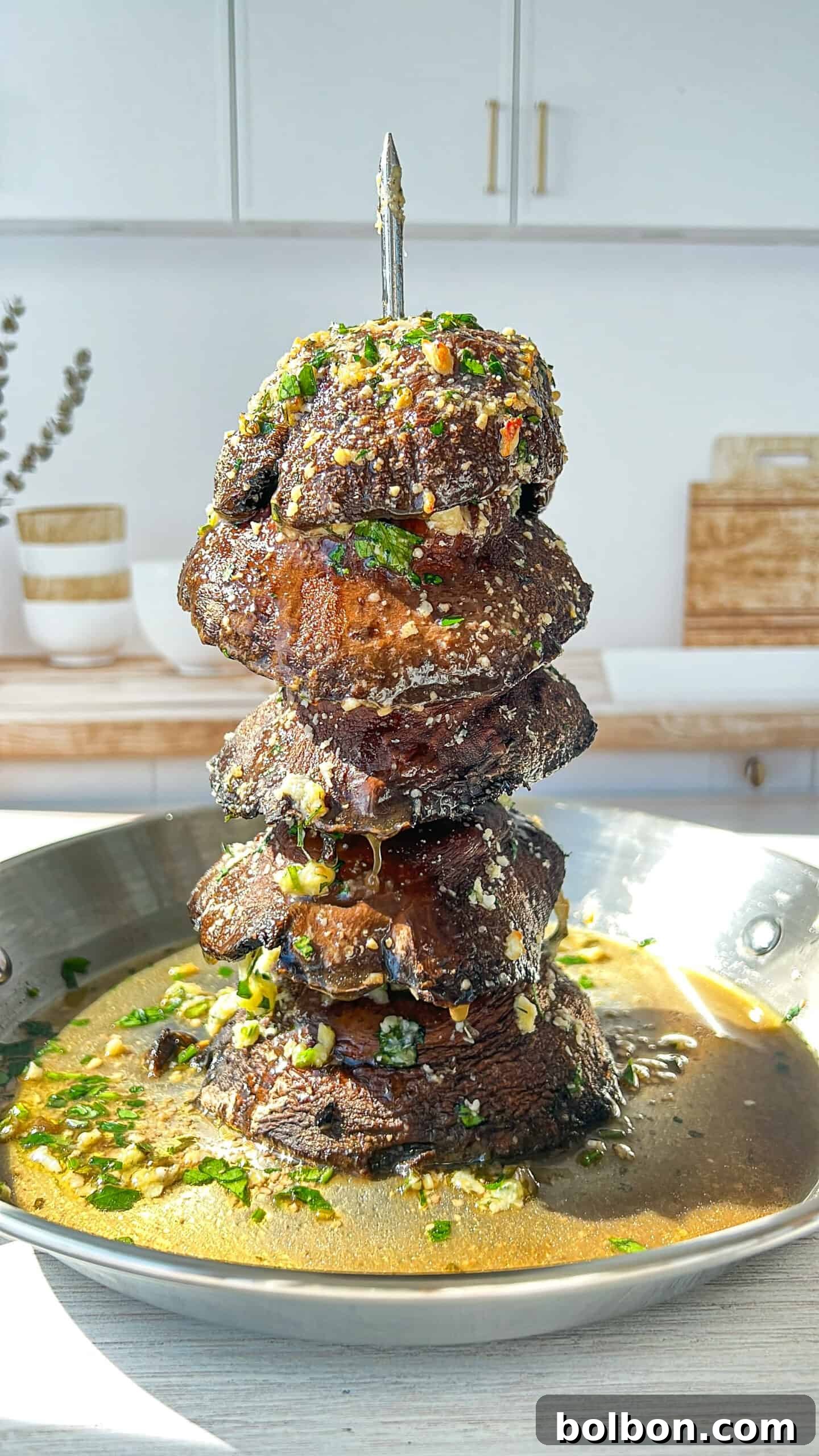 A stack of grilled meat chunks skewered vertically in a pan with sauce. The meat is garnished with herbs and grated cheese. Sunlight illuminates the dish, and the kitchen background is softly blurred.