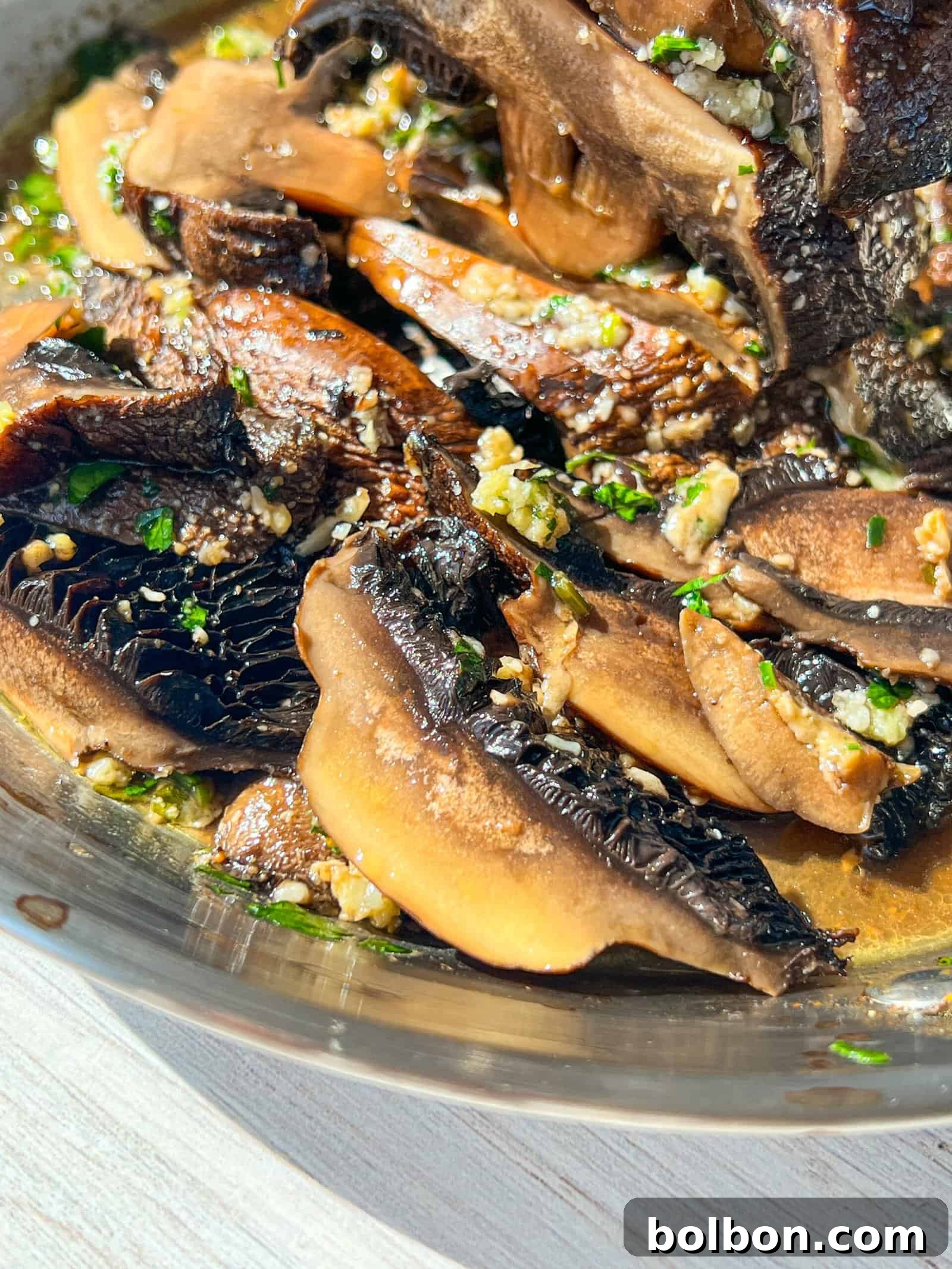 A close-up of sautéed mushrooms in a dish, glistening with olive oil and garnished with minced garlic and chopped herbs. The mushrooms are a rich brown color, and the bright sunlight enhances their texture.