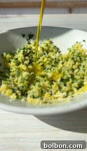 A white bowl filled with finely chopped herbs and spices is being drizzled with olive oil. The mixture includes vibrant green parsley and small white bits, possibly garlic. It's set on a light-colored wooden surface.