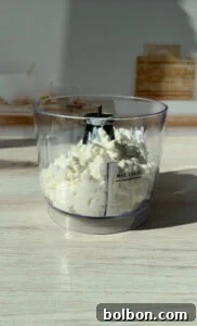 The Internet's Favorite Cottage Cheese Bagels 5 A food processor bowl filled with chopped white onions. The bowl is transparent, placed on a light wooden surface, and sunlight is casting shadows on it. The "Max Liquid" line is visible on the side.