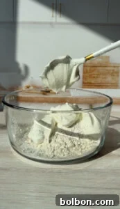 The Internet's Favorite Cottage Cheese Bagels 6 A glass bowl containing flour and scoops of cream cheese, placed on a wooden surface. A spoon above the bowl holds a dollop of cream cheese. Warm sunlight shines on the setup, with a blurred kitchen background.