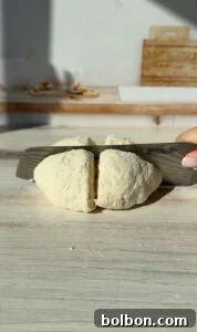 The Internet's Favorite Cottage Cheese Bagels 7 Close-up of a hand holding a knife, cutting a round, flour-covered dough on a wooden surface. Sunlight casts shadows on the table, and a kitchen counter with utensils is blurred in the background.