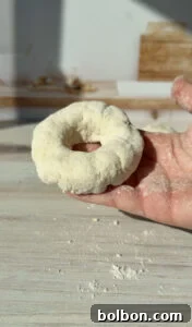 The Internet's Favorite Cottage Cheese Bagels 8 A hand holding a dough ring, dusted with flour, over a light wooden surface. The background is slightly blurred, with subtle hints of a kitchen setting, including a wooden board and some baking ingredients.