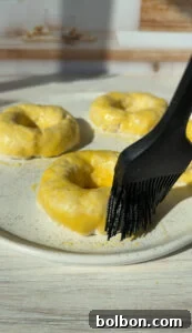 The Internet's Favorite Cottage Cheese Bagels 9 A close-up image of unbaked bagels being brushed with a shiny glaze on a white plate. Four bagels are visible, with a black brush applying the coating. The background is softly blurred, focusing on the surface texture of the dough.