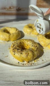 The Internet's Favorite Cottage Cheese Bagels 10 Close-up of unbaked dough rings on a plate being sprinkled with seasoning from a shaker. The dough appears soft and is on a light-colored surface, catching sunlight.