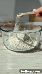 The Internet's Favorite Cottage Cheese Bagels 4 A glass bowl filled with flour on a wooden table. A hand is holding a spoon sprinkling more flour into the bowl, with the sun casting shadows and creating a warm ambiance.