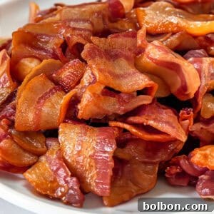 The Ultimate Guide to Crispy Bacon 3 A white plate is filled with crispy, cooked strips of bacon, arranged in a slightly scattered manner. The bacon appears glossy and has a golden-brown color, suggesting it's freshly cooked. The background is a plain, light surface.