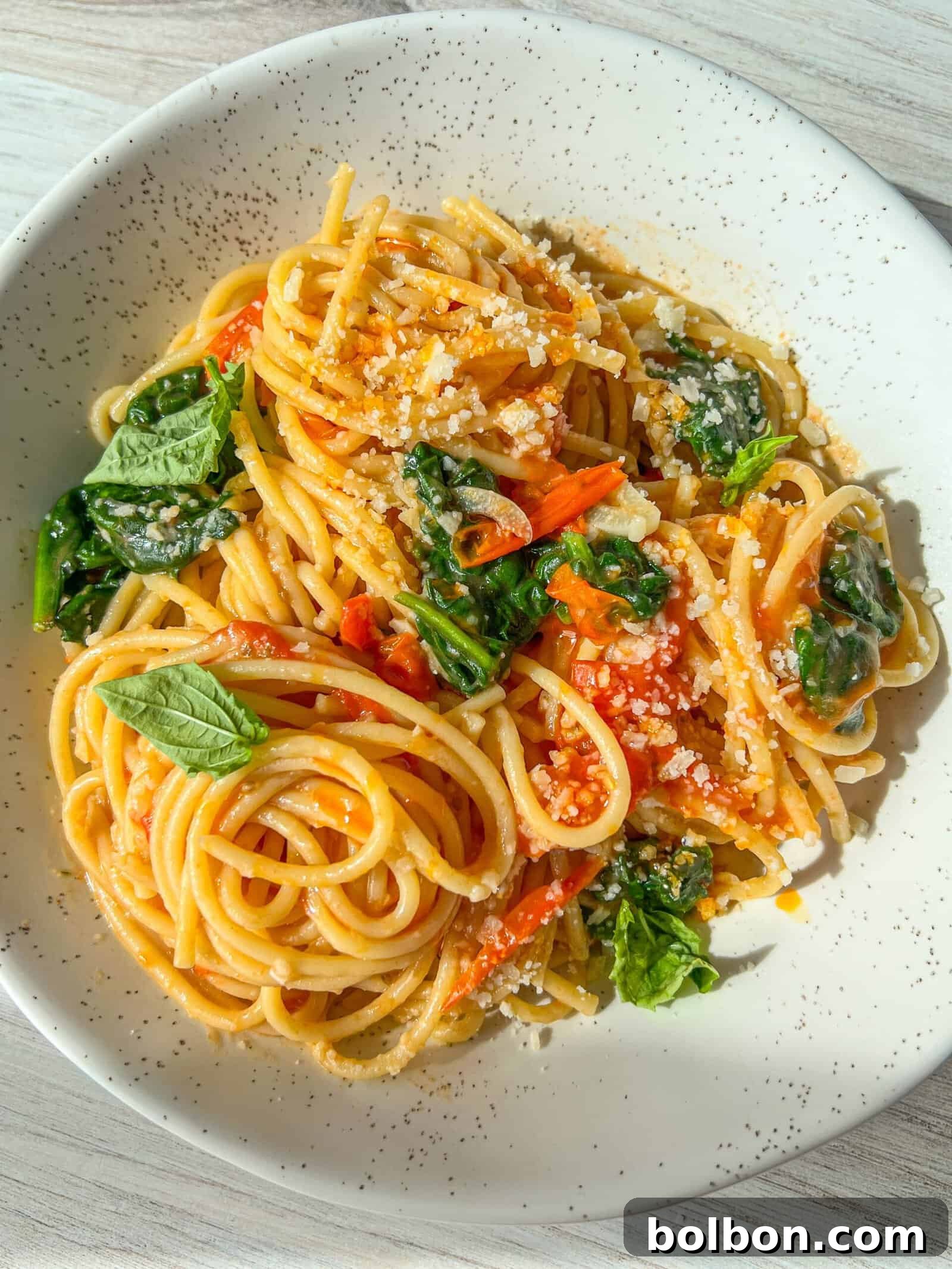 Meghan Markle's One-Pot Pasta with Spinach and Tomatoes A plate of spaghetti pasta topped with vibrant spinach leaves, diced tomatoes, and sprinkled Parmesan cheese. The dish is arranged on a white, speckled plate, set on a light wooden surface, highlighting the fresh ingredients and the simple elegance of the meal.