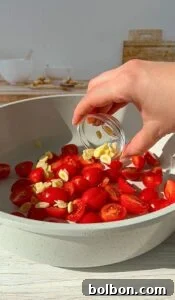Meghan Markle's Royal Cake Recipe 2 A hand is sprinkling sliced garlic from a small glass dish into a bowl filled with halved cherry tomatoes. In the background, there are bowls and a cutting board on a light wood surface.