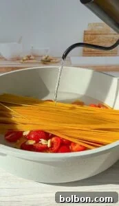 Meghan Markle's Royal Cake Recipe 3 A bowl of uncooked spaghetti, halved cherry tomatoes, and garlic cloves is being filled with hot water from a kettle, set against a kitchen background.
