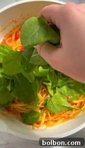 Meghan Markle's Royal Cake Recipe 4 A hand adding fresh spinach to a pot of spaghetti with tomato sauce. The bright green spinach contrasts with the red sauce and yellow pasta, creating a vibrant and colorful dish.