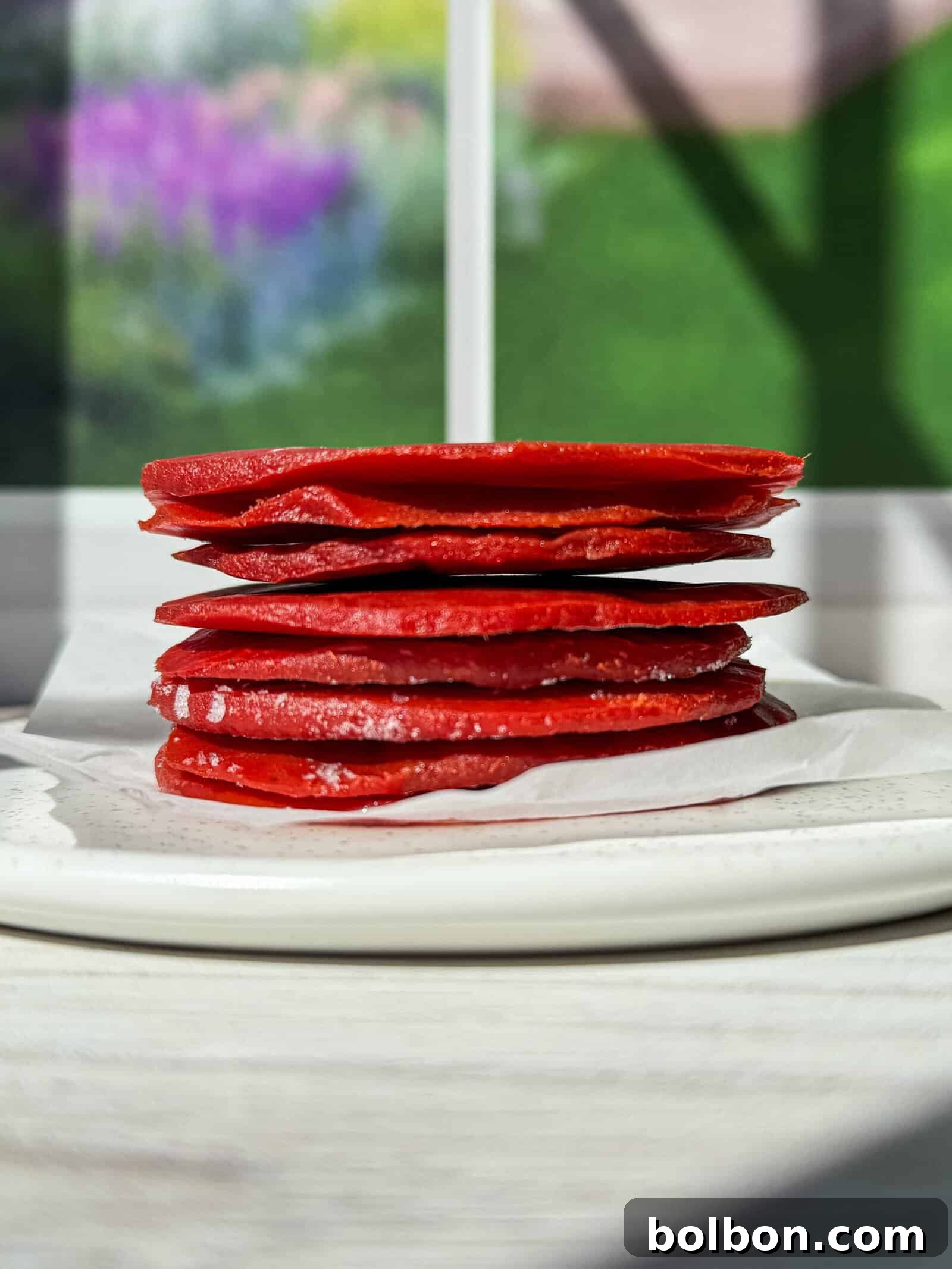 Genius Tomato Paste Tips 2 A stack of thin, red fruit leather squares on a white plate with parchment paper. The background shows a blurred view of a window with green trees and a colorful garden outside. Sunlight casts shadows across the table.