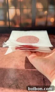 Genius Tomato Paste Tips 6 A hand holding a stack of white napkins, each with a round red stain in the center. The background is softly blurred with a grid-like window pattern, casting a warm light.