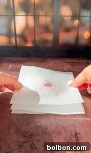 Genius Tomato Paste Tips 4 Two hands hold a stack of thin tissue papers with a small brownish-red stain in the center. The background features a window with a blurred, warm view of trees and sunlight.