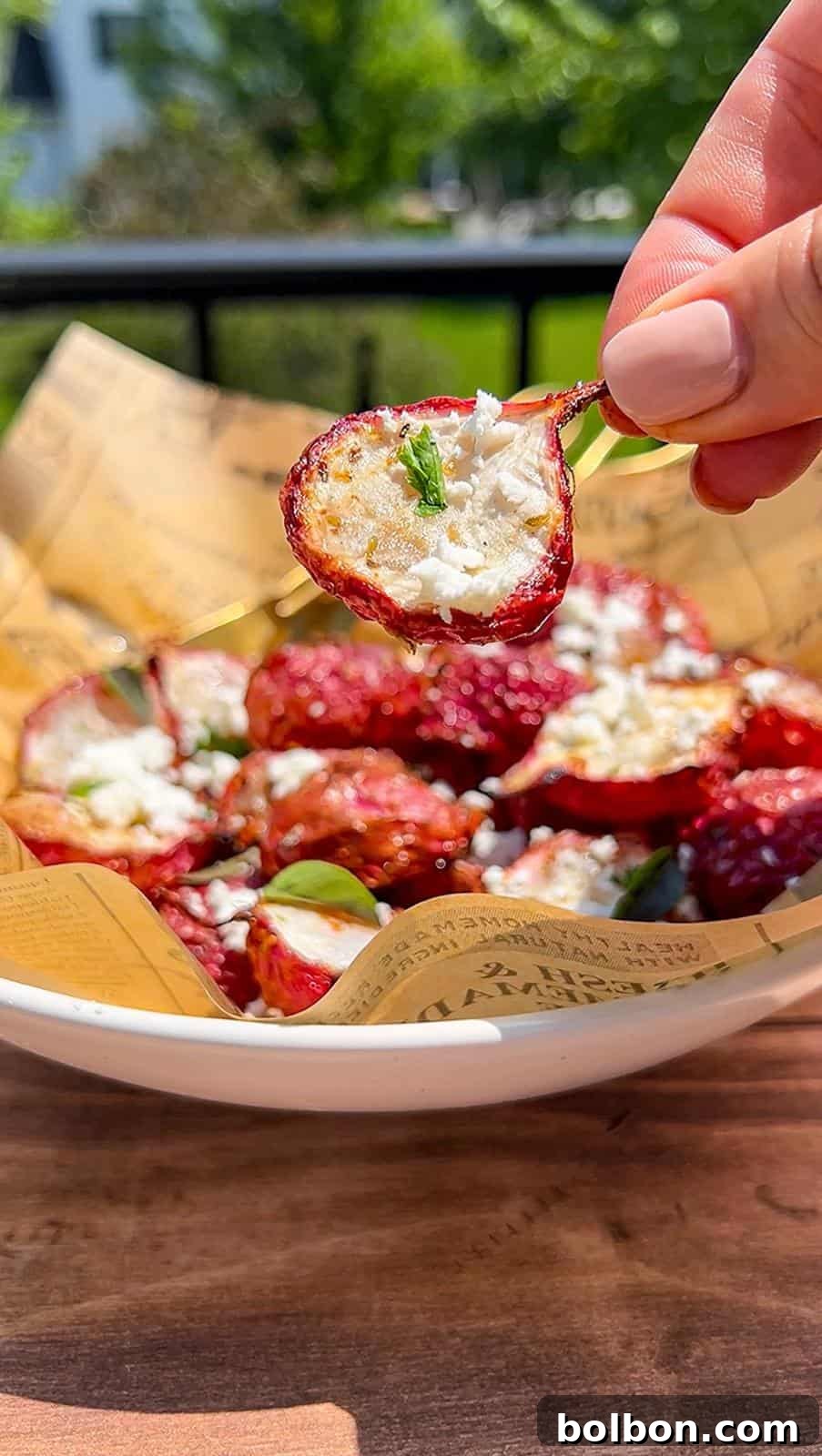 A hand holds a roasted air fryer radish topped with crumbled cheese and herbs above a bowl filled with similar tomatoes, all resting on printed parchment paper, with greenery visible in the background.