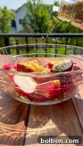 A glass bowl filled with halved radishes, lemon zest, and olive oil sits on a wooden table outdoors, while dried herbs are being sprinkled from a jar above the bowl. Greenery and a house appear in the blurred background.