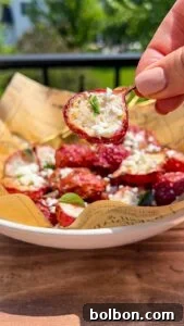 A hand holds a roasted cherry tomato topped with crumbled cheese and herbs above a bowl filled with similar tomatoes, all resting on printed parchment paper, with greenery visible in the background.