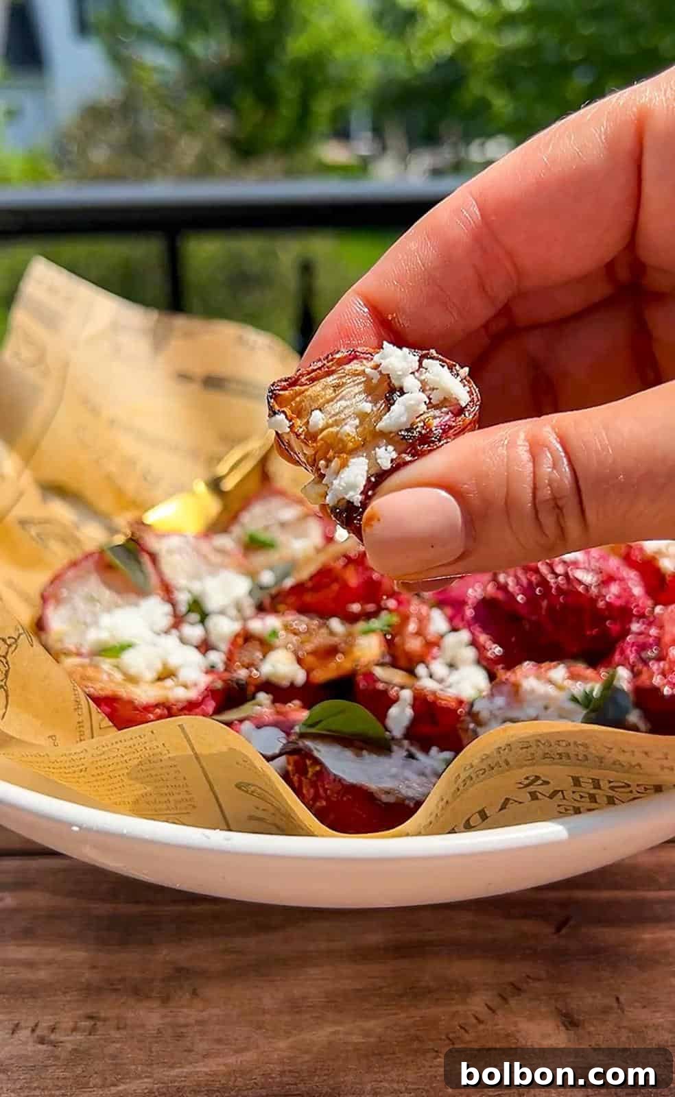 A hand holding a sliced roasted air fryer radish topped with crumbled cheese over a bowl of roasted beets garnished with cheese and herbs, served on paper-lined dish outdoors.