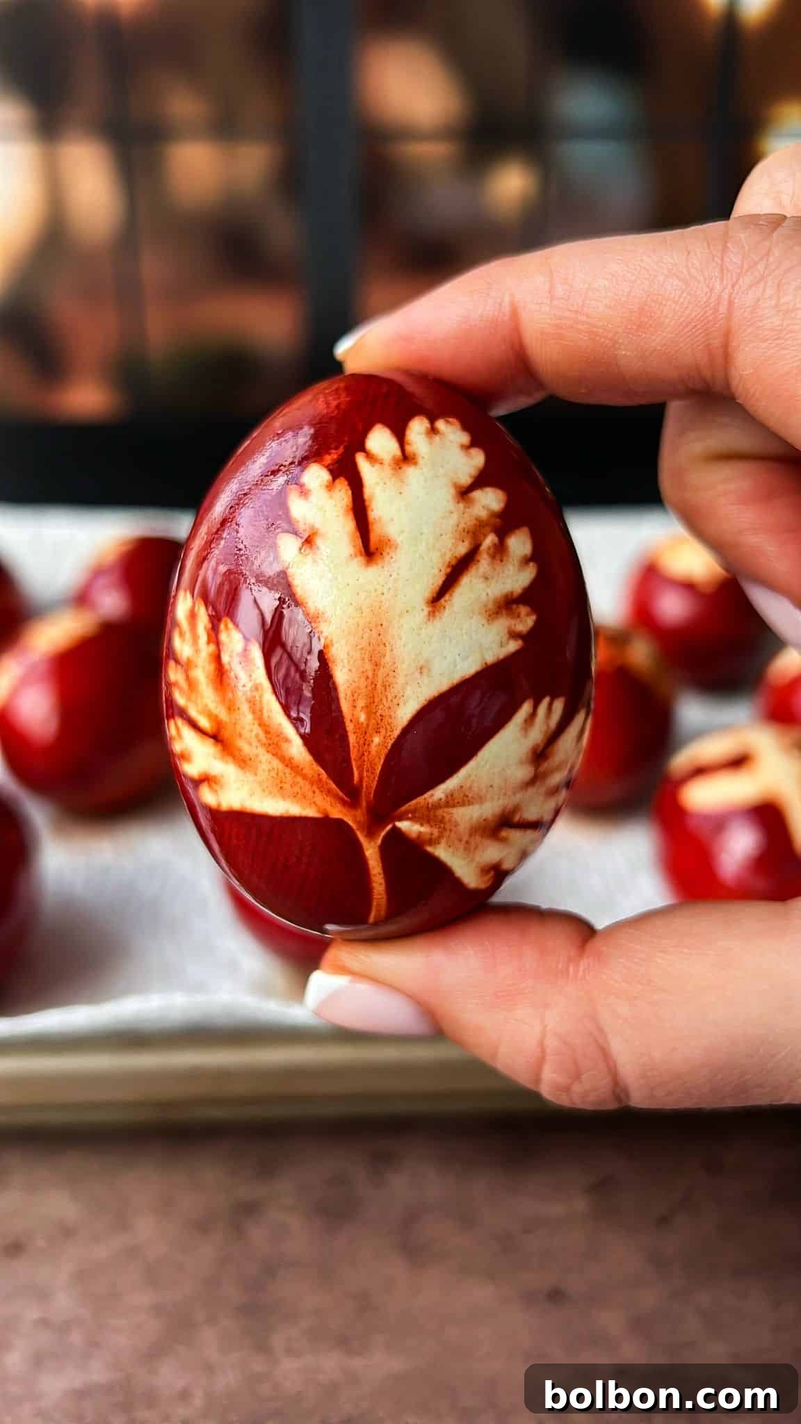 Doğanın Renkleriyle Paskalya Yumurtaları 2 A hand holding a deep red Easter egg featuring a leaf pattern in beige. The background shows other similarly decorated eggs on a tray.