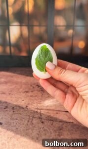Doğanın Renkleriyle Paskalya Yumurtaları 6 A hand holding a white egg with a green leaf on it, set against a warm, softly blurred background with grid-like window patterns and a rustic brown surface.