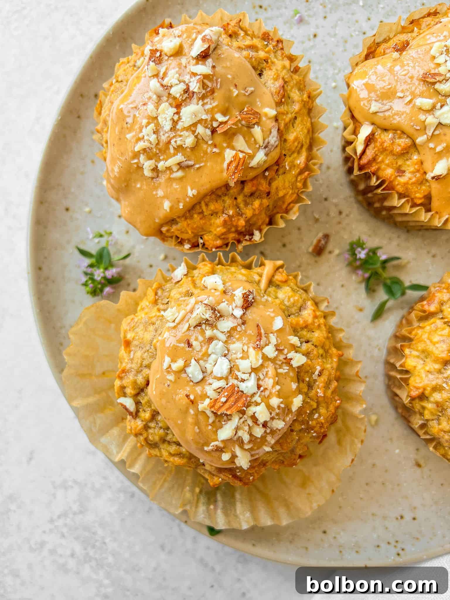 Rise and Shine Muffins 2 Three healthy breakfast muffins topped with a light spread and chopped nuts sit invitingly on a speckled ceramic plate. Each muffin is nestled in its crinkled paper liner, and delicate small purple flowers are scattered artfully on the plate, enhancing the rustic and natural presentation. The background features a soft, light gray surface, providing a clean and appealing contrast to the vibrant muffins.