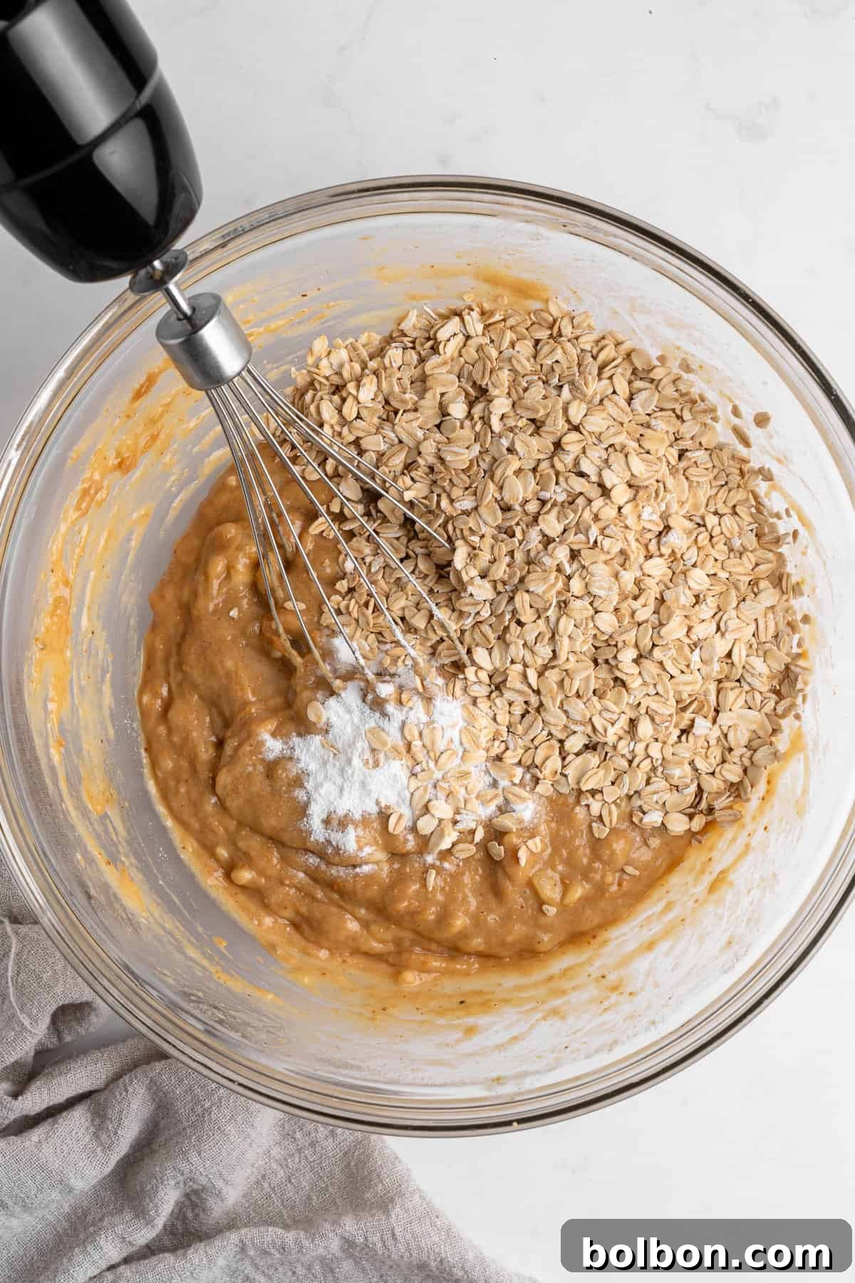 Rise and Shine Muffins 5 Inside a clear glass bowl, a delicious mixture of batter with mashed bananas and oats is being expertly mixed with a whisk, showing the baking powder visibly sprinkled on top. The bowl is positioned on a sleek marble countertop, next to a cozy beige cloth, creating an inviting kitchen scene.