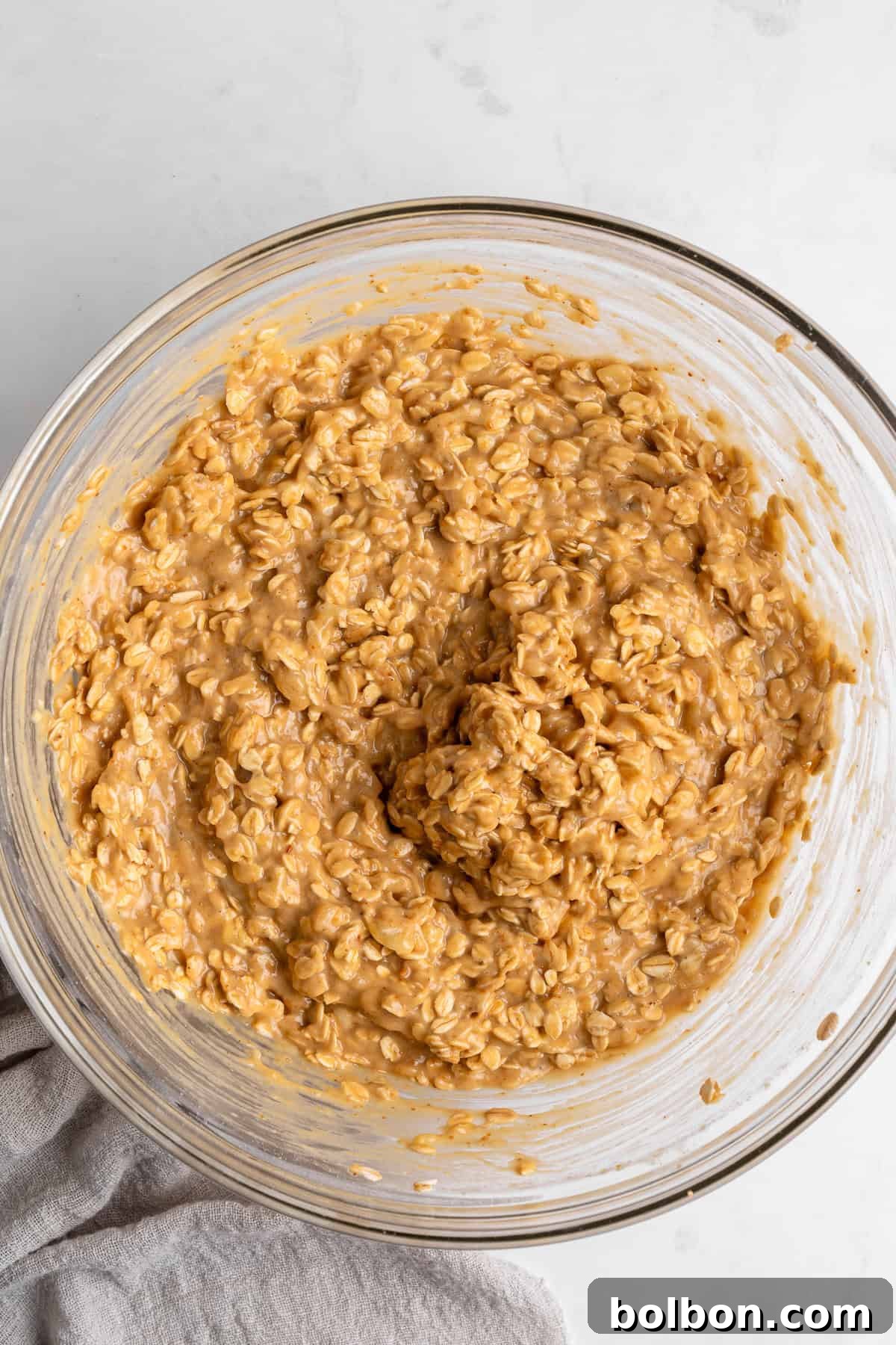 Rise and Shine Muffins 6 A glass bowl filled with a creamy peanut butter oatmeal mixture, perfectly blended and ready for baking, sits gracefully on a pristine white surface. A charming beige cloth napkin peeks out partially from the bottom left corner, adding a subtle touch of elegance to the kitchen setup.