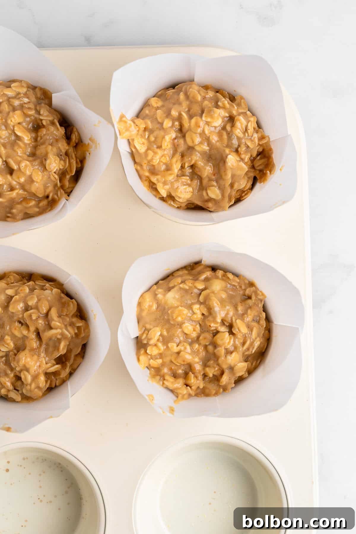 Rise and Shine Muffins 7 Four healthy breakfast muffins, generously filled with a thick, chunky peanut butter oat batter, are neatly placed within the individual cups of a white muffin tray. Two of the cups are full of the rich batter, while the other two compartments remain empty, showcasing a light gray surface beneath.
