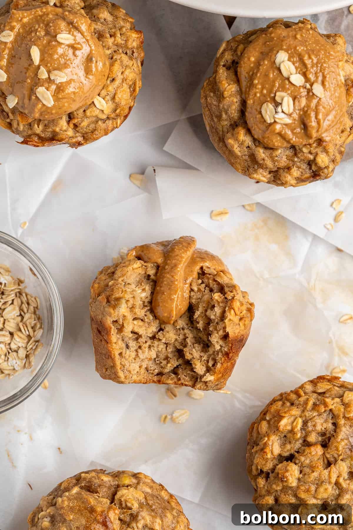 Rise and Shine Muffins 8 Healthy Breakfast Muffins, artfully topped with creamy peanut butter and scattered oats, are arranged on delicate parchment paper, hinting at their wholesome ingredients. One muffin has been temptingly bitten, revealing a moist and tender interior, inviting a taste. A small glass bowl filled with rolled oats sits nearby, perfectly complementing the rustic and natural presentation.