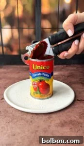 A hand uses a can opener to open a Unico Tomato Paste can labeled with "Italian Herbs." The can is placed on a white plate against a blurred background.