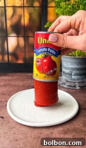 A hand holding an upside-down can of tomato paste, with the paste standing upright on a white plate. A green plant and a black window grid are in the background.