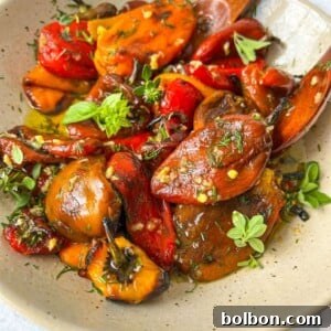 A beige bowl filled with roasted tomatoes, eggplants, and roasted bell peppers garnished with fresh herbs sits on a white surface, with a wooden spoon resting in the bowl.