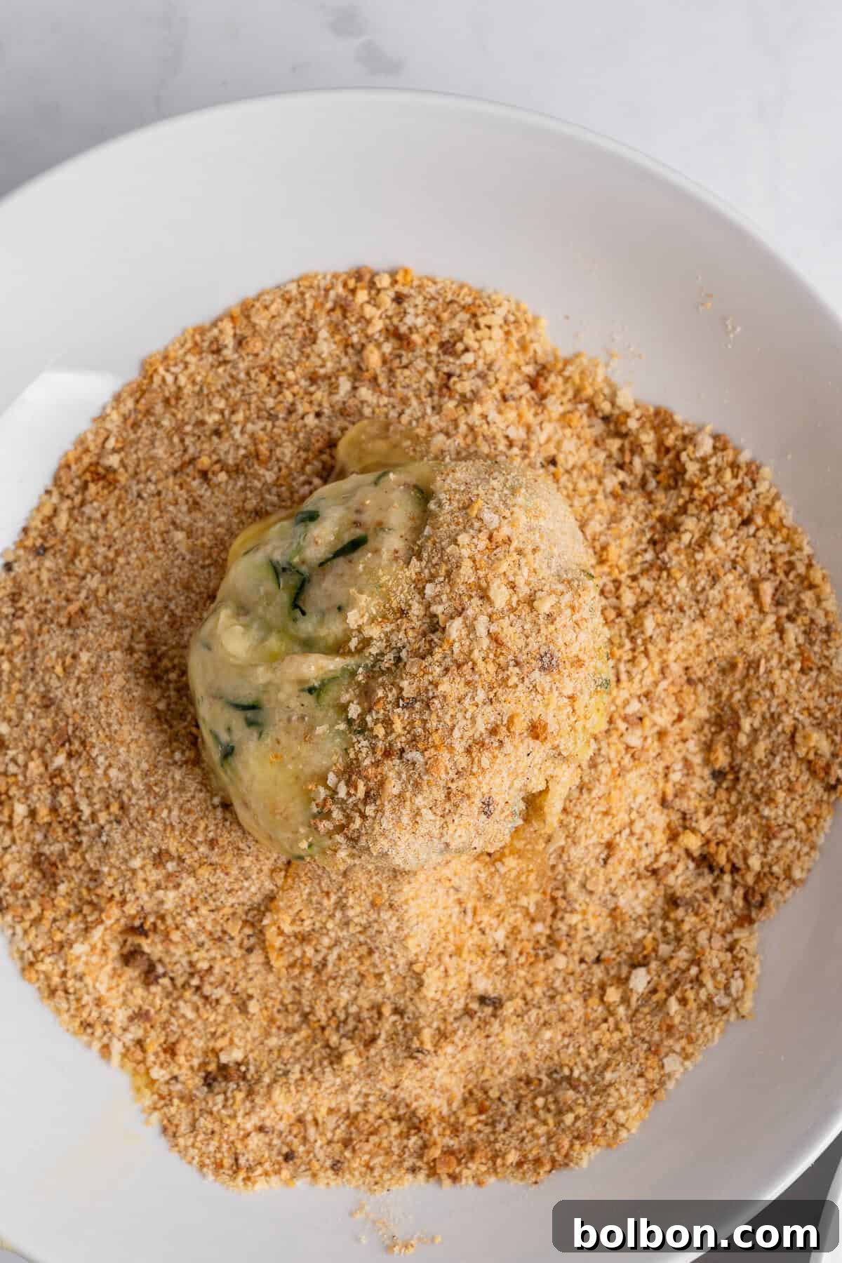 Crispy Zucchini Fritters 11 A perfectly formed ball of uncooked dough, rich with visible herbs and spices, is expertly coated in a fine layer of breadcrumbs on a pristine white plate, set against a classic marble surface. This image captures the crucial step of preparing food for frying, ensuring a crispy outer texture.