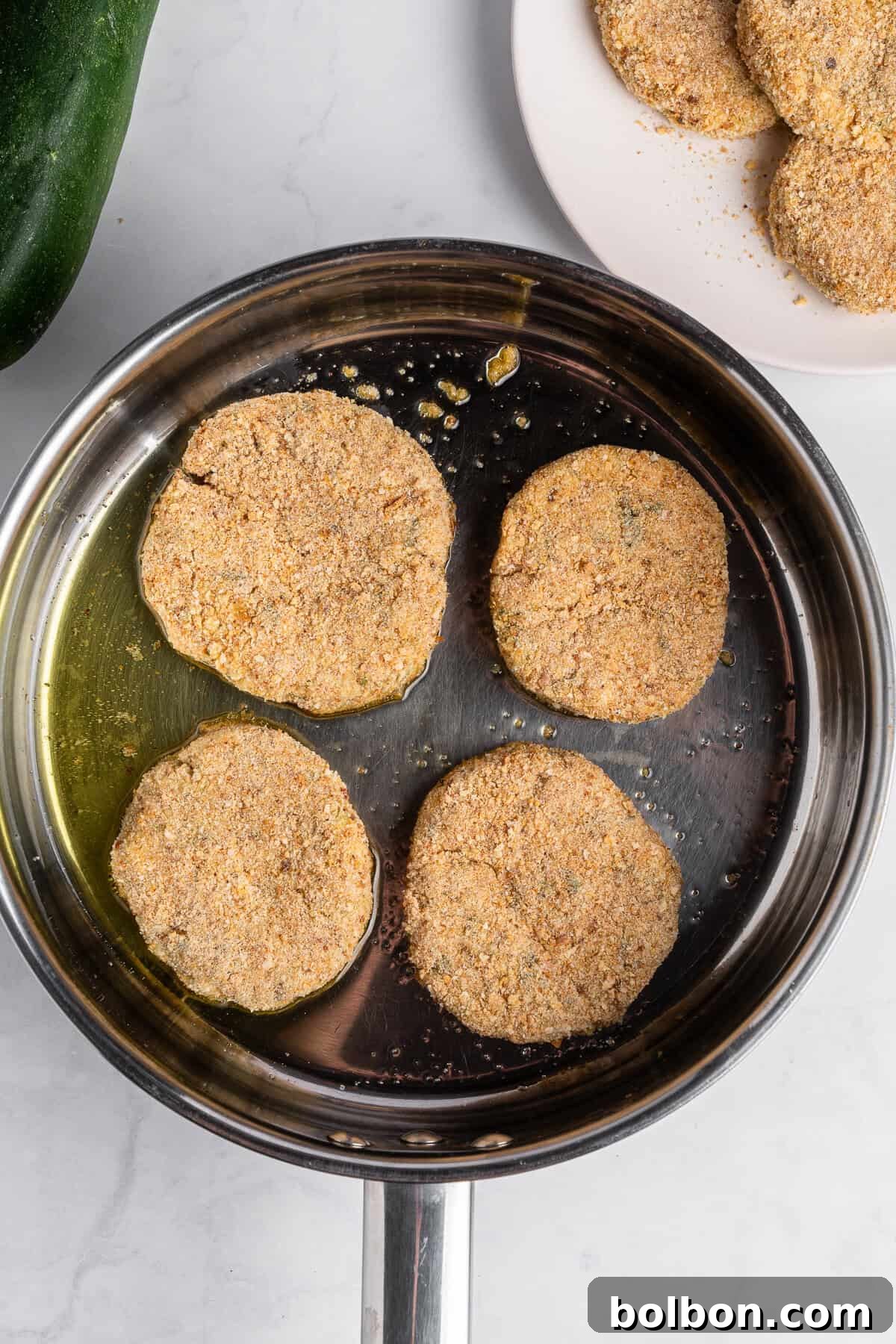 Crispy Zucchini Fritters 12 Four perfectly breaded zucchini slices are actively frying in a gleaming stainless steel pan, surrounded by hot oil, showcasing the sizzle and transformation. In the background, a plate with more uncooked, breaded slices awaits its turn, indicating a batch cooking process.