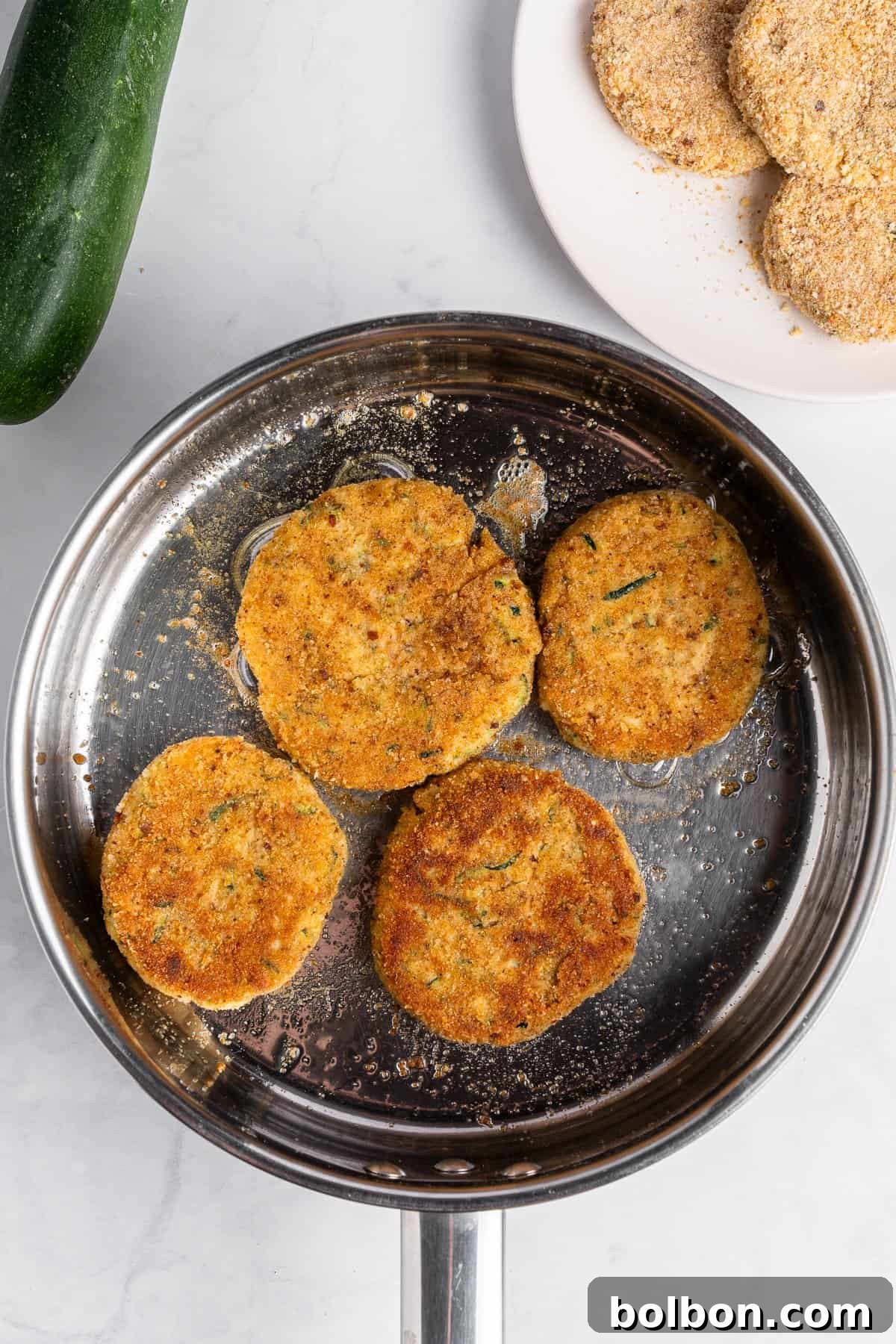 Crispy Zucchini Fritters 13 Four golden-brown zucchini fritters are beautifully frying in a stainless steel pan, their crispy edges promising a delightful texture. A fresh zucchini is positioned nearby, and a plate featuring breadcrumbs alongside uncooked fritters is visible in the background, all resting on a white marble surface, highlighting the various stages of preparation.