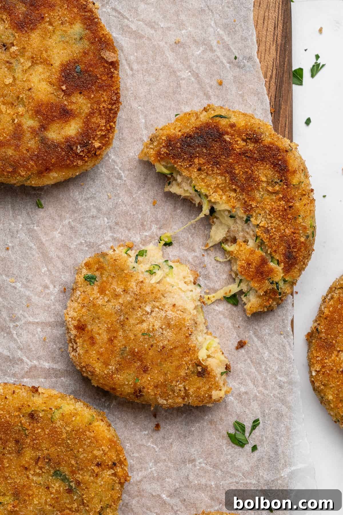 Crispy Zucchini Fritters 14 Top view of several golden-brown, breaded, and fried patties arranged on parchment paper, ready to be served. One patty is broken in half, revealing a creamy, herb-flecked filling, emphasizing its tender interior. Other patties remain whole and are garnished with a sprinkle of chopped parsley, adding a fresh green touch.