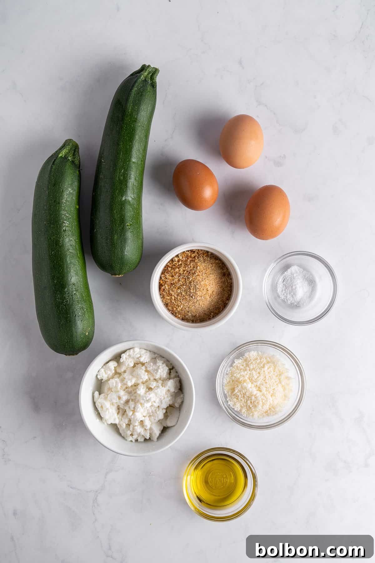 Crispy Zucchini Fritters 3 Ingredients for zucchini cakes arranged on a marble surface: two whole zucchinis, three eggs, breadcrumbs in a bowl, salt in a small bowl, ricotta cheese in a bowl, grated Parmesan in a bowl, and a bowl of olive oil, ready for preparation.