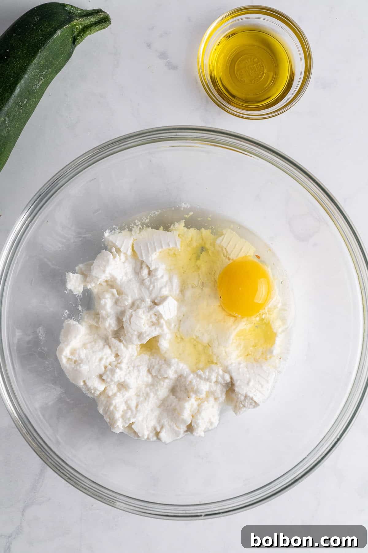 Crispy Zucchini Fritters 4 A clear glass bowl filled with creamy ricotta cheese sits on a marble countertop, with a cracked egg beside it, its yolk and white ready to be incorporated into the mixture. A small glass bowl of olive oil and a fresh zucchini are nearby, setting the scene for cooking.