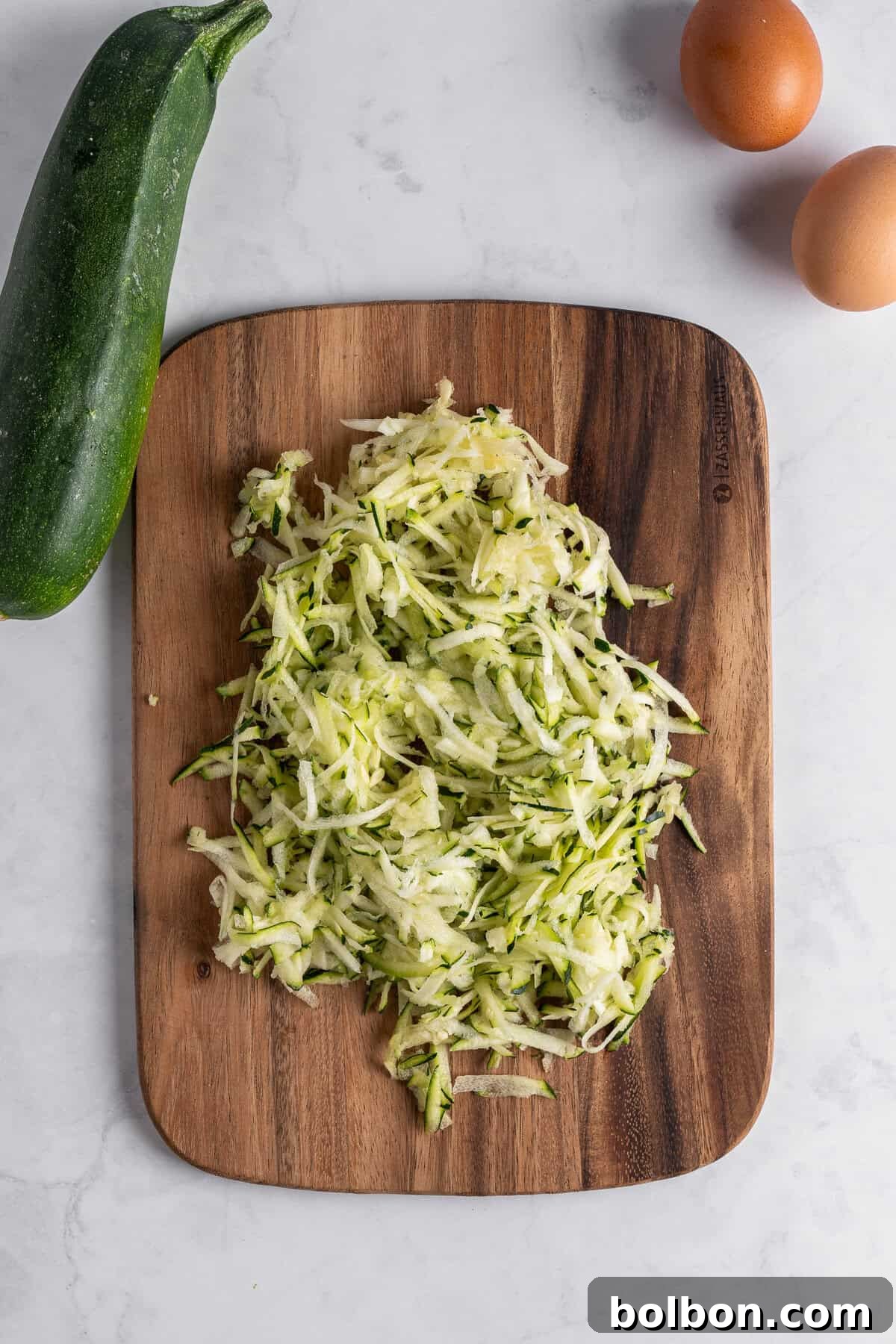 Crispy Zucchini Fritters 5 A wooden cutting board holds a generous pile of finely shredded zucchini, ready for squeezing out excess moisture. A whole, fresh zucchini is placed beside the board, along with two brown eggs, all arranged on a pristine white marble surface, highlighting the fresh ingredients.