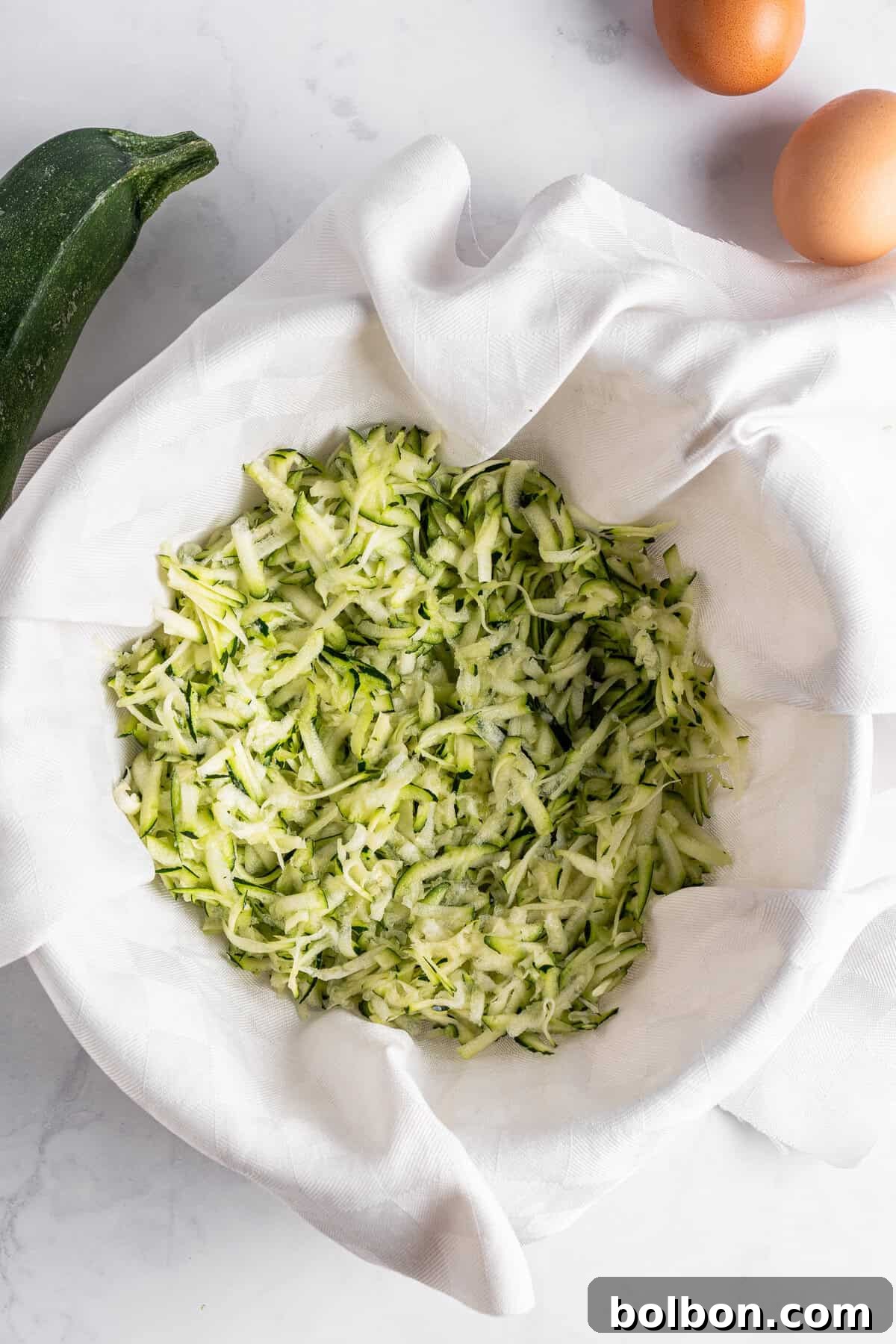 Crispy Zucchini Fritters 6 A clear glass bowl is lined with a clean white cloth, holding a fresh pile of grated zucchini on a bright marble countertop. A whole zucchini and two brown eggs are positioned nearby, emphasizing the freshness of the ingredients.