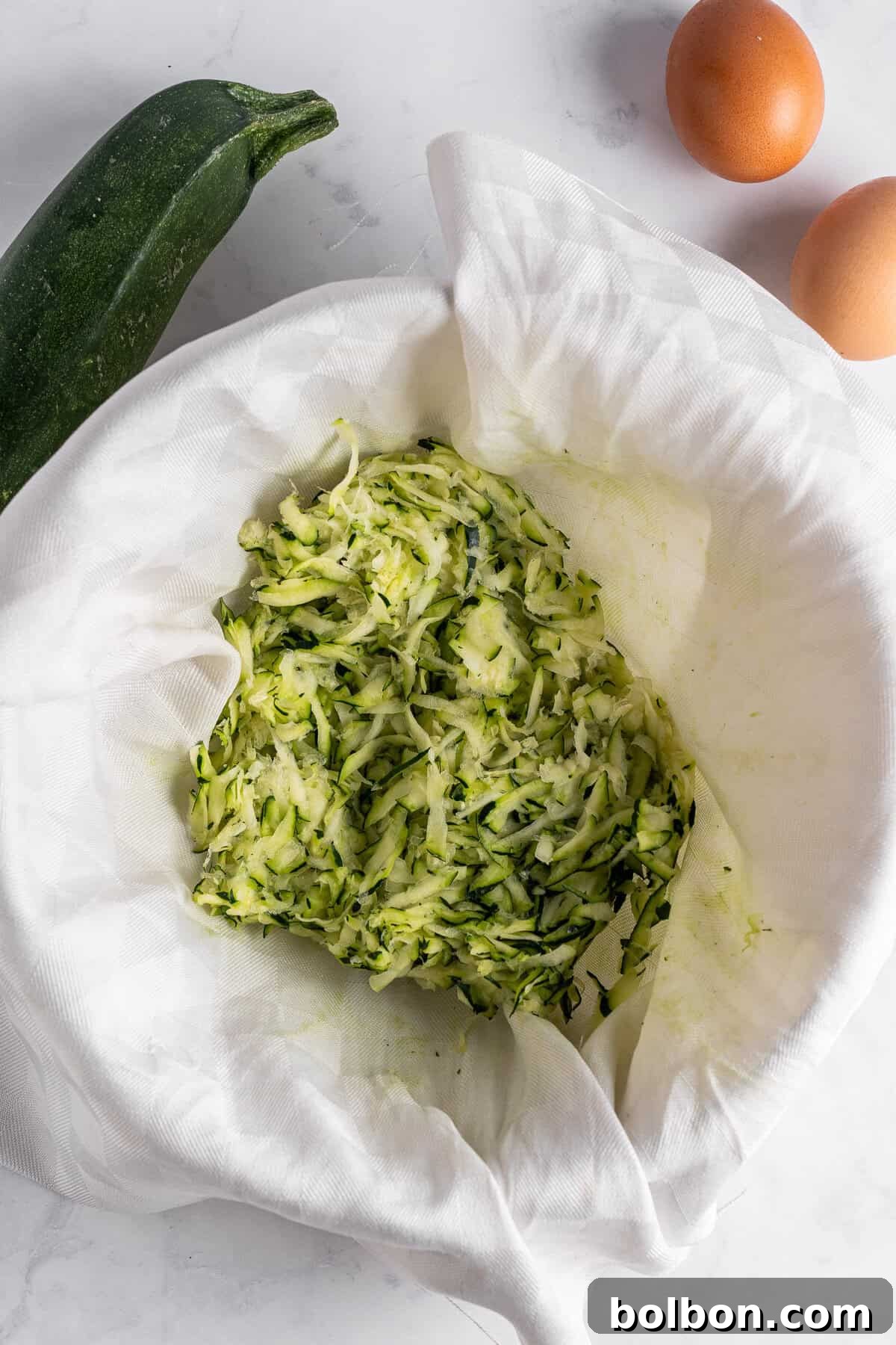 Crispy Zucchini Fritters 7 A pristine white cloth holds a generous pile of shredded zucchini, with visible green and white strands, ready for squeezing. A whole zucchini and two brown eggs are carefully placed on the white marble countertop nearby, highlighting the preparation process.