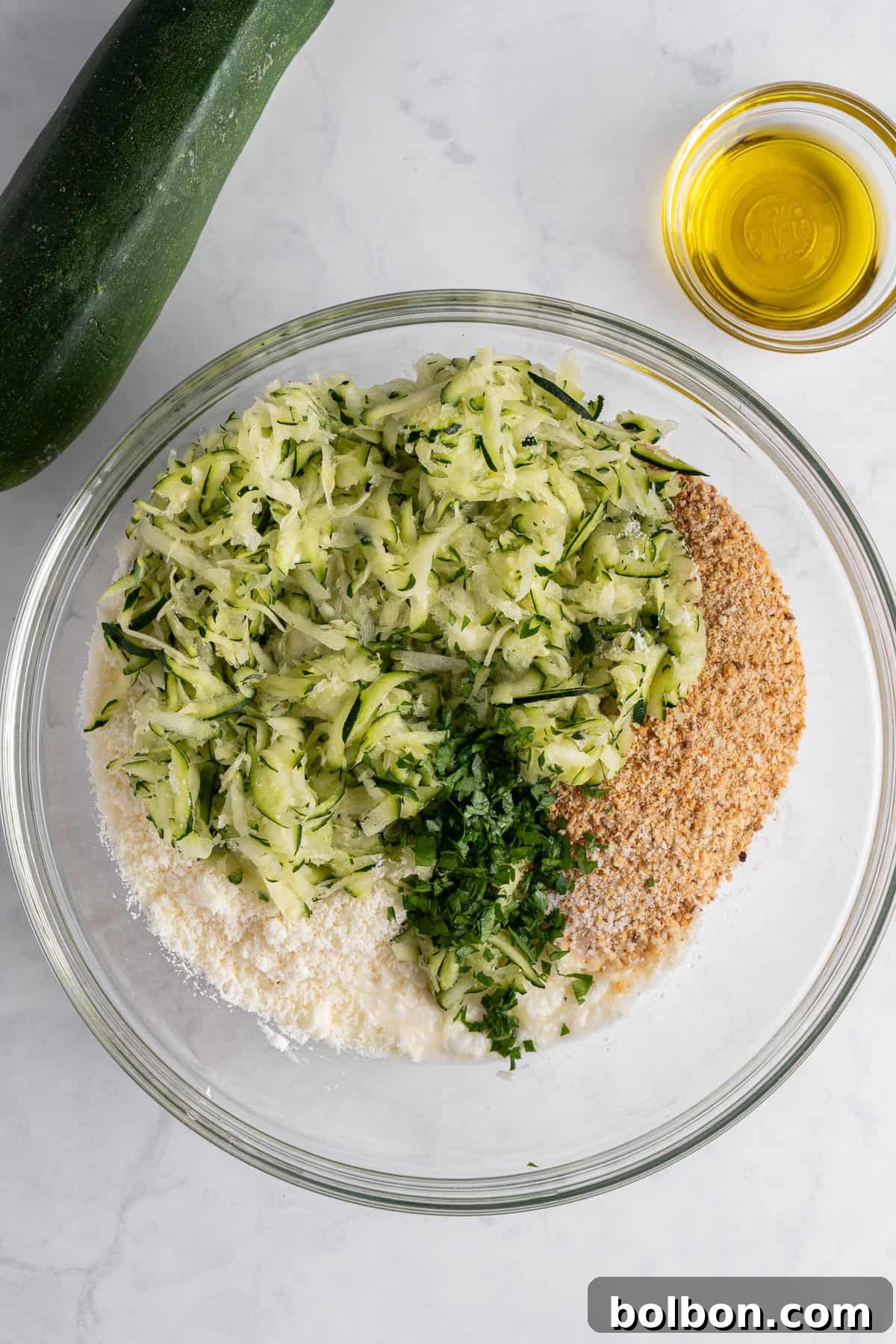 Crispy Zucchini Fritters 8 A large mixing bowl on a marble surface contains grated zucchini, Italian breadcrumbs, grated Parmesan cheese, and finely chopped fresh herbs. A whole zucchini and a small bowl of olive oil are positioned adjacent to the bowl, ready for the next steps in the recipe.