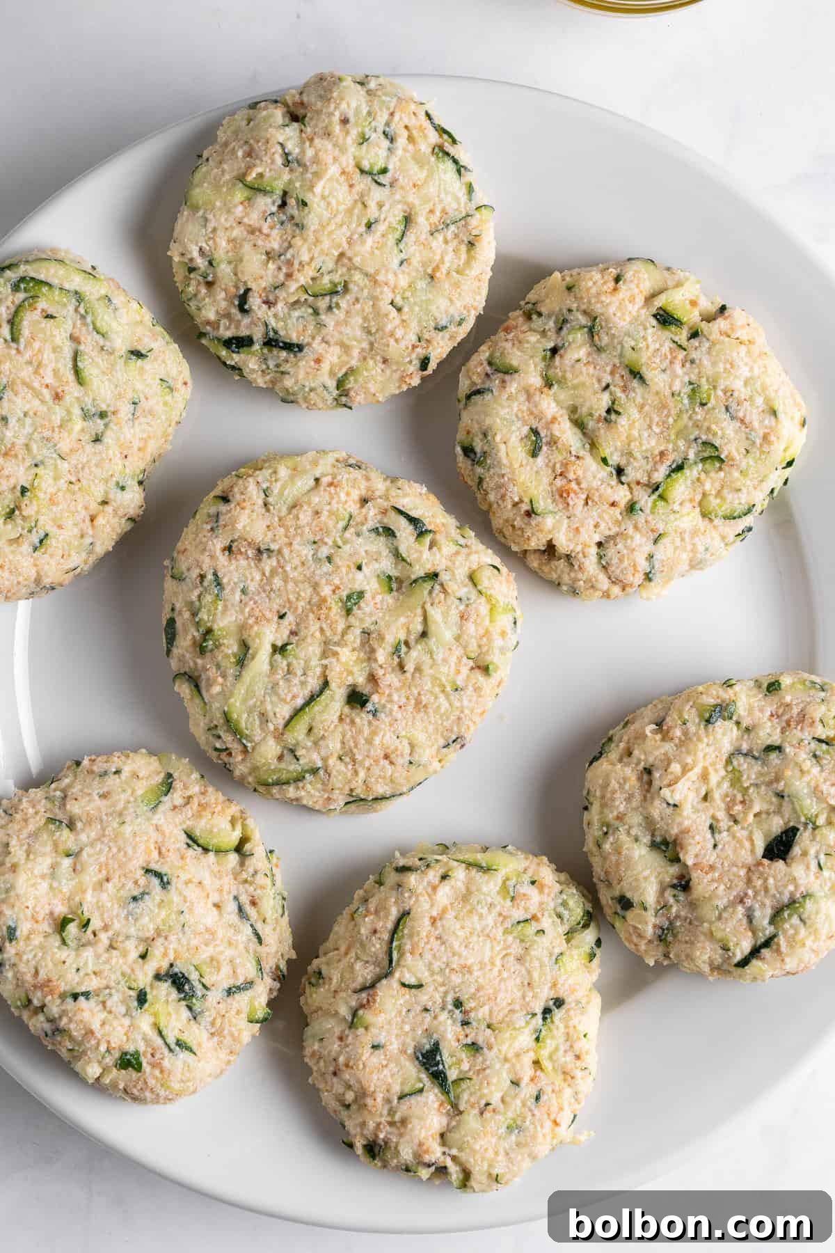 Crispy Zucchini Fritters 9 A white plate displays six uncooked, perfectly round patties made from a mixture of grated zucchini and breadcrumbs. The patties exhibit a speckled texture with visible green bits of zucchini, suggesting their fresh ingredients and readiness for cooking.