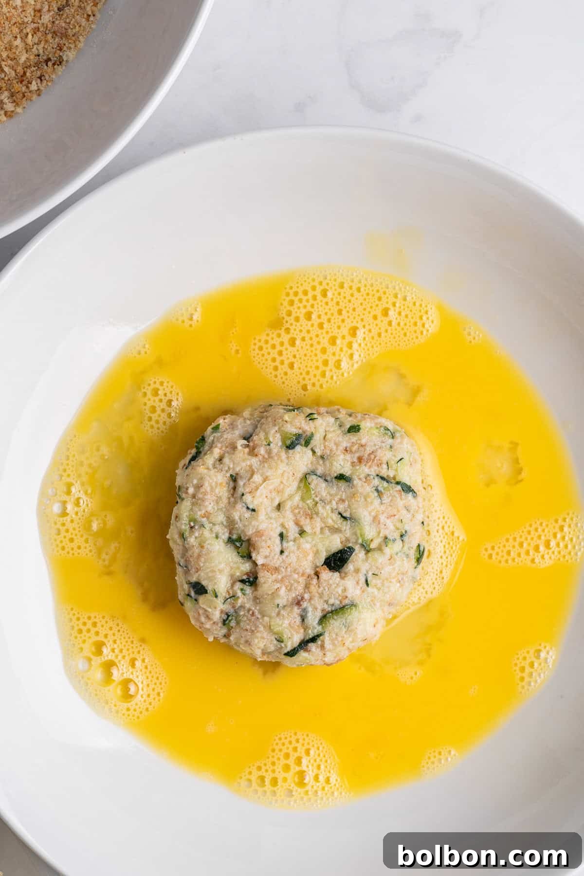 Crispy Zucchini Fritters 10 A pristine white plate holds a savory mixture, possibly minced meat or vegetables with herbs, surrounded by a pool of beaten egg. The egg yolk and whites are thoroughly mixed, creating a golden liquid that partially coats the formed patty, ready for breading.