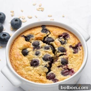 A baked oat dish topped with blueberries, served in a white ramekin. The surface is golden brown with bursts of juicy blueberries. Some oats and fresh blueberries are scattered on the white background.