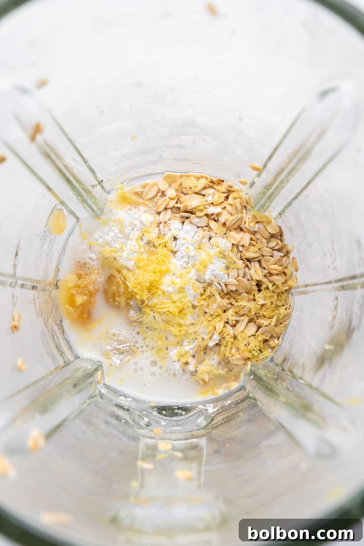 Wholesome Blueberry Oat Loaf 4 A blender contains oats, lemon zest, milk, and banana. The ingredients are ready to be blended, with oats and lemon zest scattered on top, and milk partially covering them. The mixture appears in a glass blender jug.