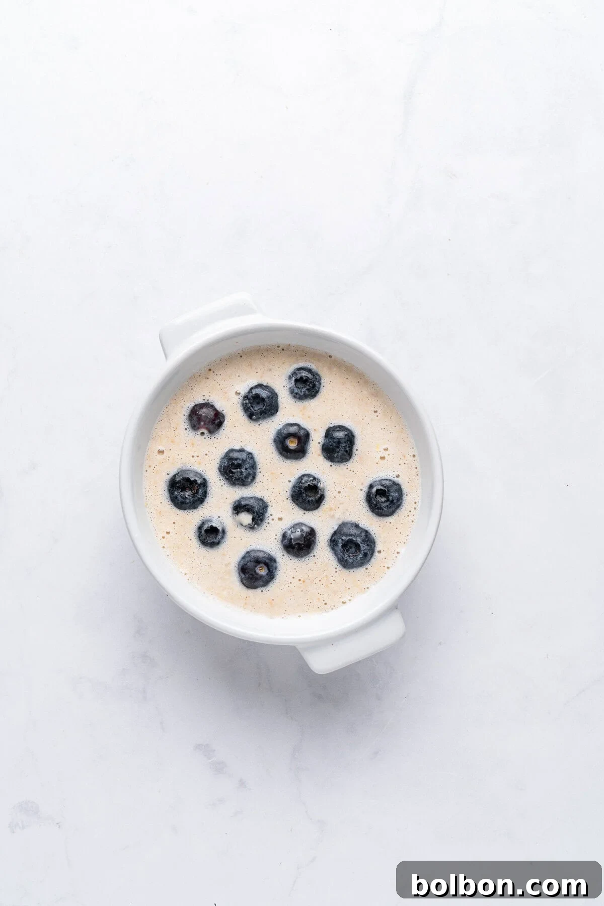 Wholesome Blueberry Oat Loaf 5 A white bowl filled with a creamy dish topped with scattered blueberries, set on a light-colored marble surface.