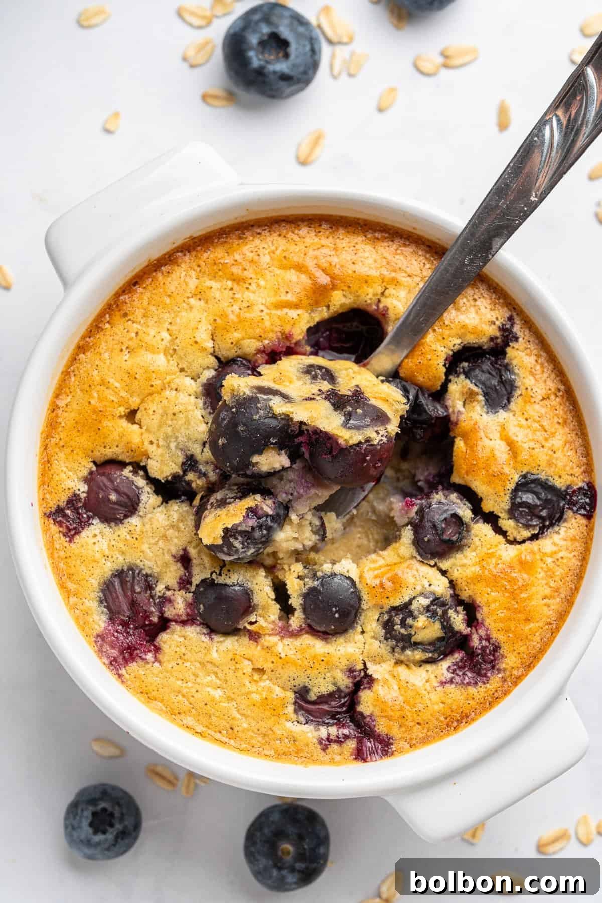 Wholesome Blueberry Oat Loaf 6 A bowl of baked oatmeal with blueberries on top and scattered around, with a spoon scooping a portion. The dish is set on a light surface with a few oat flakes nearby, creating a cozy breakfast scene.